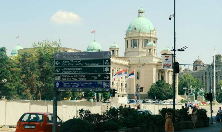 Skupština Srbije tokom sunčanog dana prema kojoj ide crveni automobil.