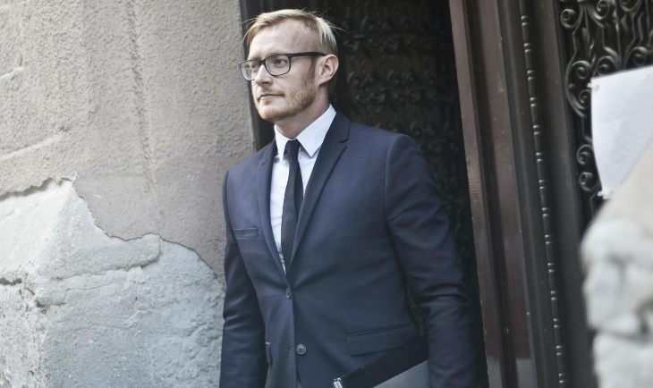 Čovek sa naočarima za vid u teget odelu drži laptop i izlazi iz zgrade
