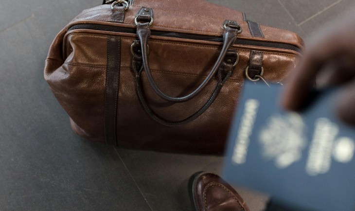 Kožna putna torba braon boje na podu pored čoveka koji drži pasoš