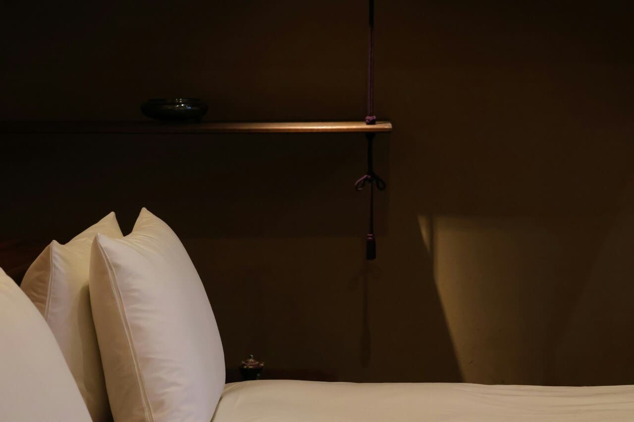 Beli jastuci na krevetu pored tamnog zida sa drvenom policom i visećim ukrasom