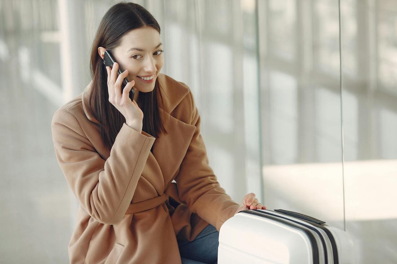 Žena u bež kaputu sedi pored belog kofera i razgovara telefonom