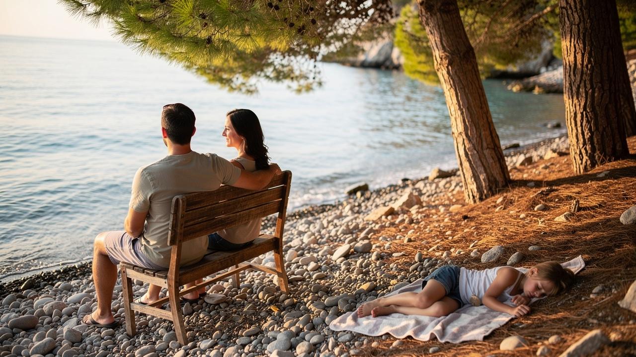Par sedi na klupi pored mora dok dete spava na peškiru ispod borova